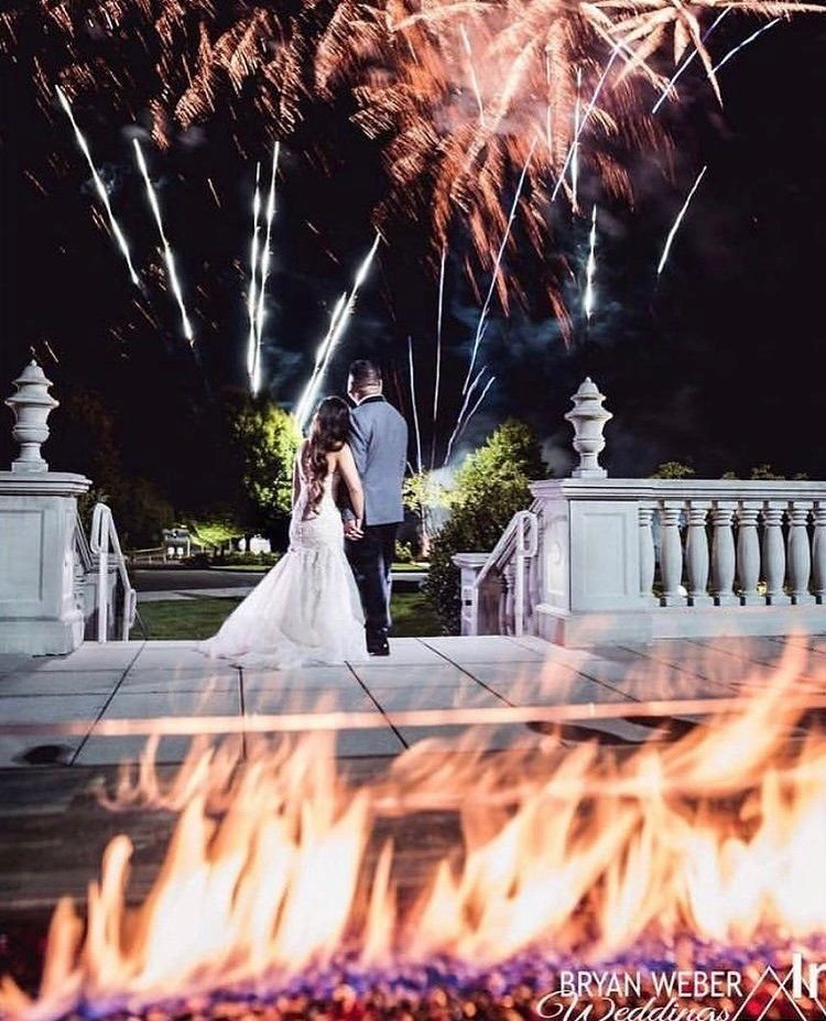 Newlyweds on a stone terrace at night watching aerial fireworks burst above, with a low gas flame in the foreground