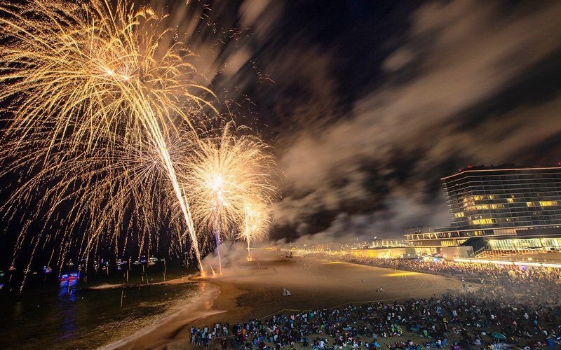 Large gold fireworks bursting above a beachfront crowd at night, with a hotel and shoreline lights at right