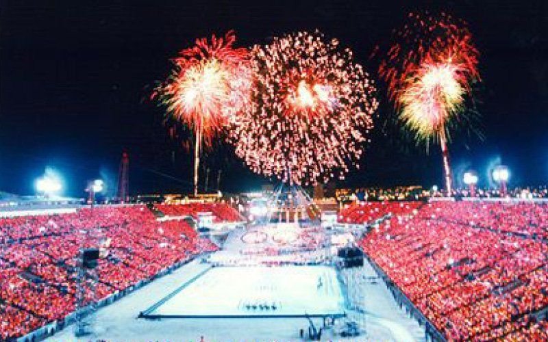 Red, white and pink fireworks over a packed stadium at night, with a lit field and a crowd in red