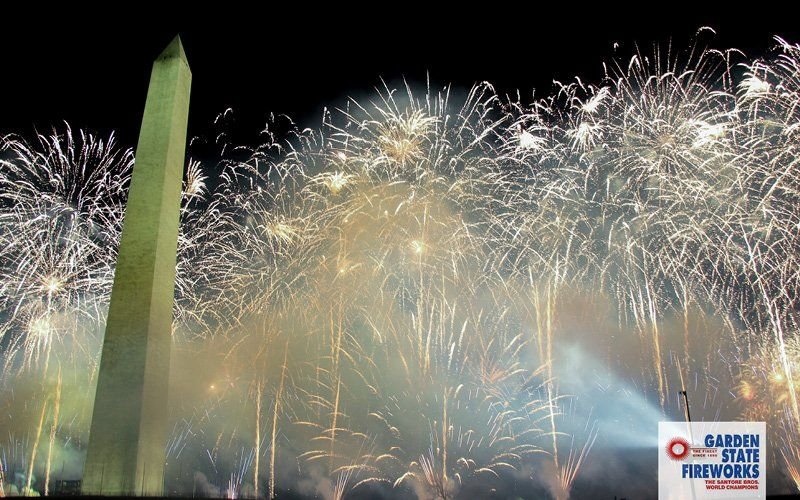 White and gold fireworks filling the sky beside the Washington Monument, with smoke drifting across the Mall