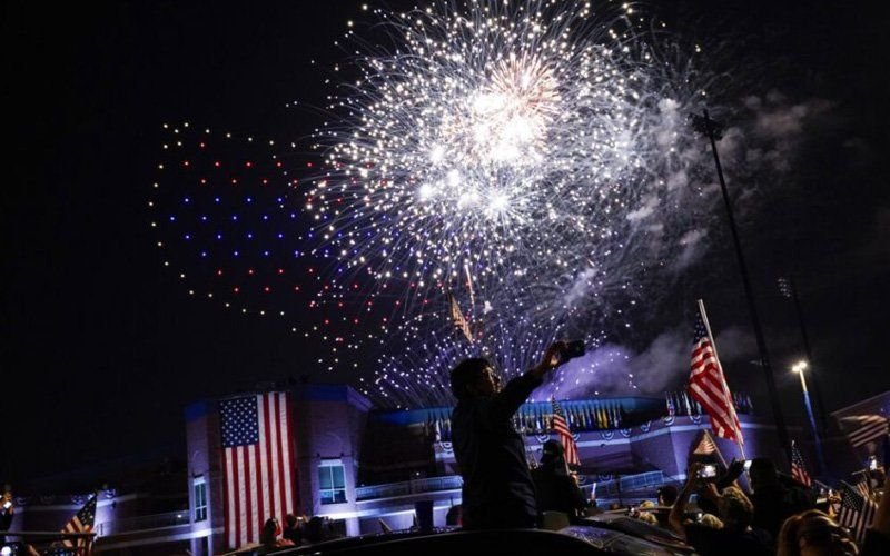 Spectators with flags watching white fireworks and a red and blue drone light formation over a lit building