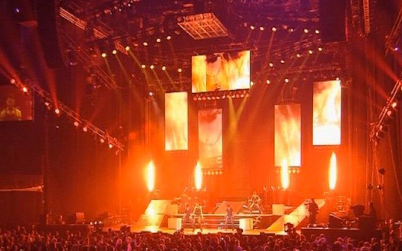 Flame jets lining a concert stage in front of tall video screens, with a packed crowd in the foreground