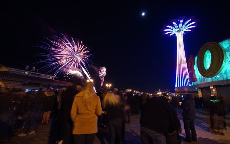 Crowd on a boardwalk watching fireworks at night near a lit, tall amusement ride