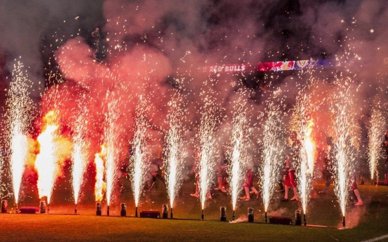 Rows of fountain sparklers and flame jets along a stadium field as players enter through smoke
