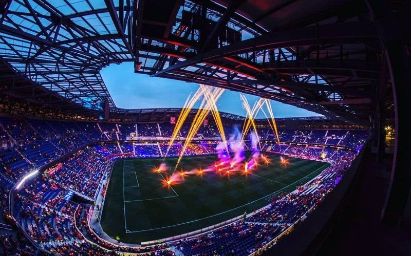 Wide view inside a roofed soccer stadium as gold comets and pink pyrotechnics fire above the field