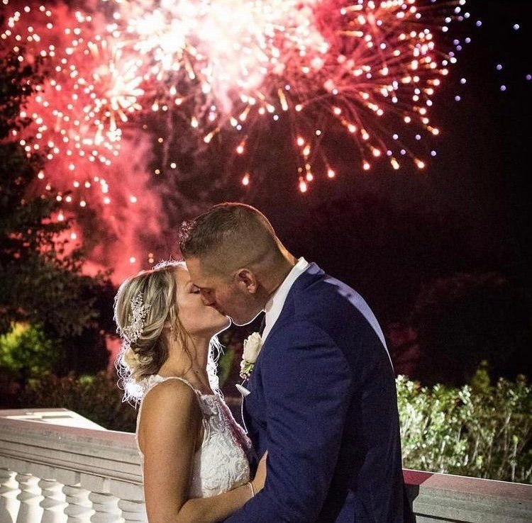 Bride and groom kissing on a stone terrace at night as pink and red fireworks burst overhead