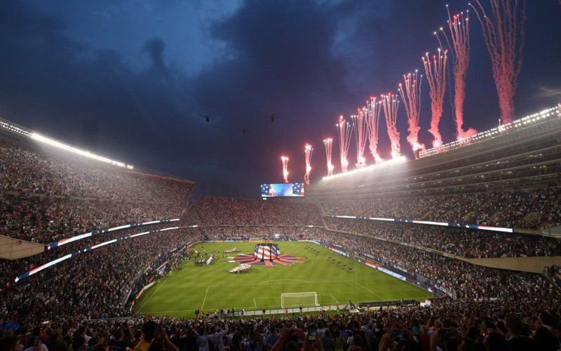 Pink and red fireworks and comets rising from the stands of a packed stadium at dusk, with a large flag on the field
