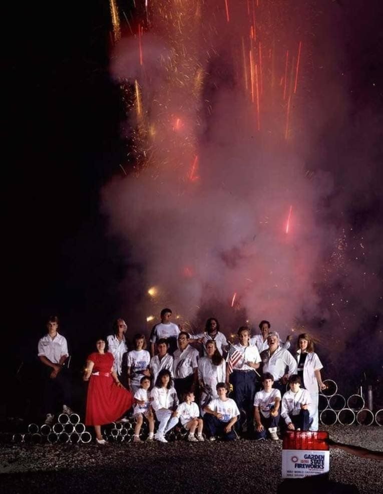 Family and crew in white shirts posed at night in front of a glowing cloud of firework smoke and sparks