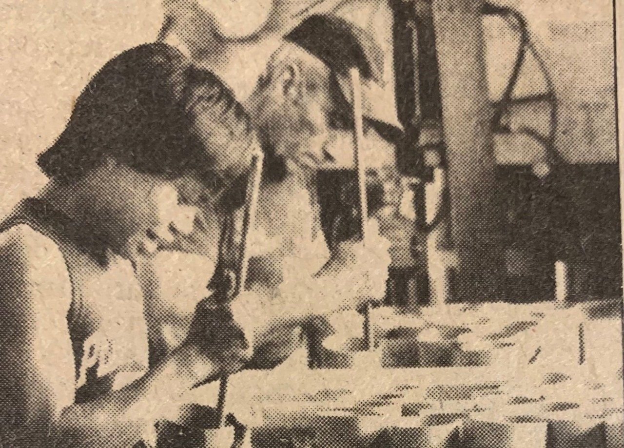 Two workers filling rows of small tubes with long tools at a workbench in a 1986 newspaper print