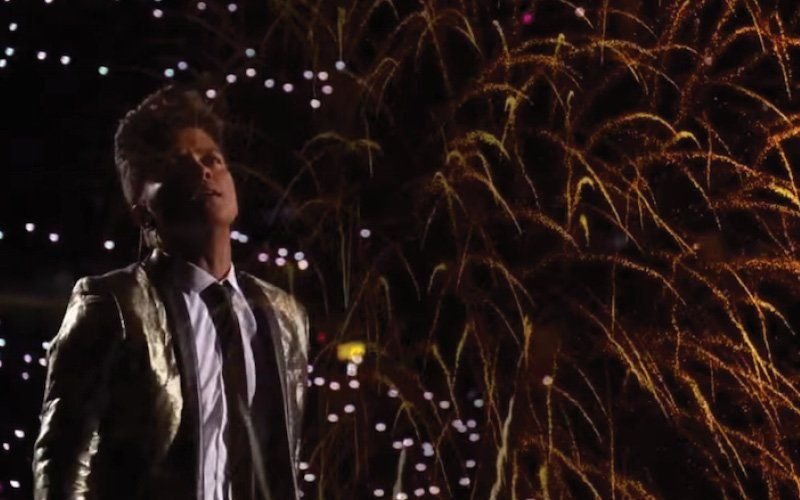 Performer in a sequined jacket looking up as gold fireworks fall behind him in a darkened stadium