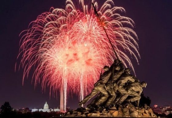 Red and gold fireworks bursting behind the Iwo Jima Memorial statue, with the Capitol dome in the distance